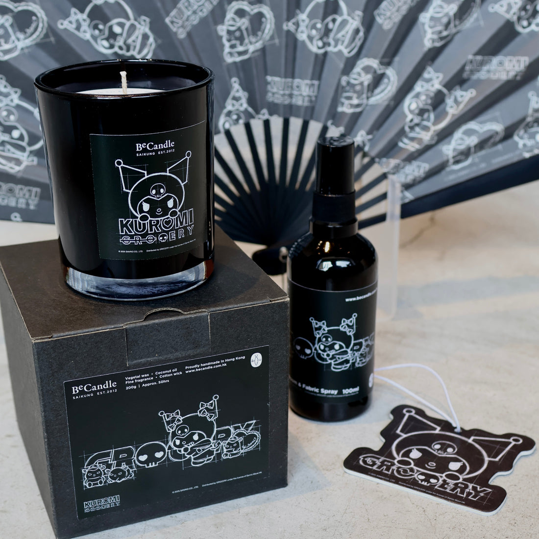 Black Kuromi candle and bottle with cat design on a gray surface, fan in the background
