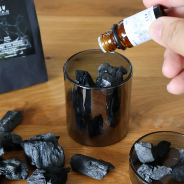 Person adding liquid from a bottle into a glass container with charcoal pieces on a wooden surface.
